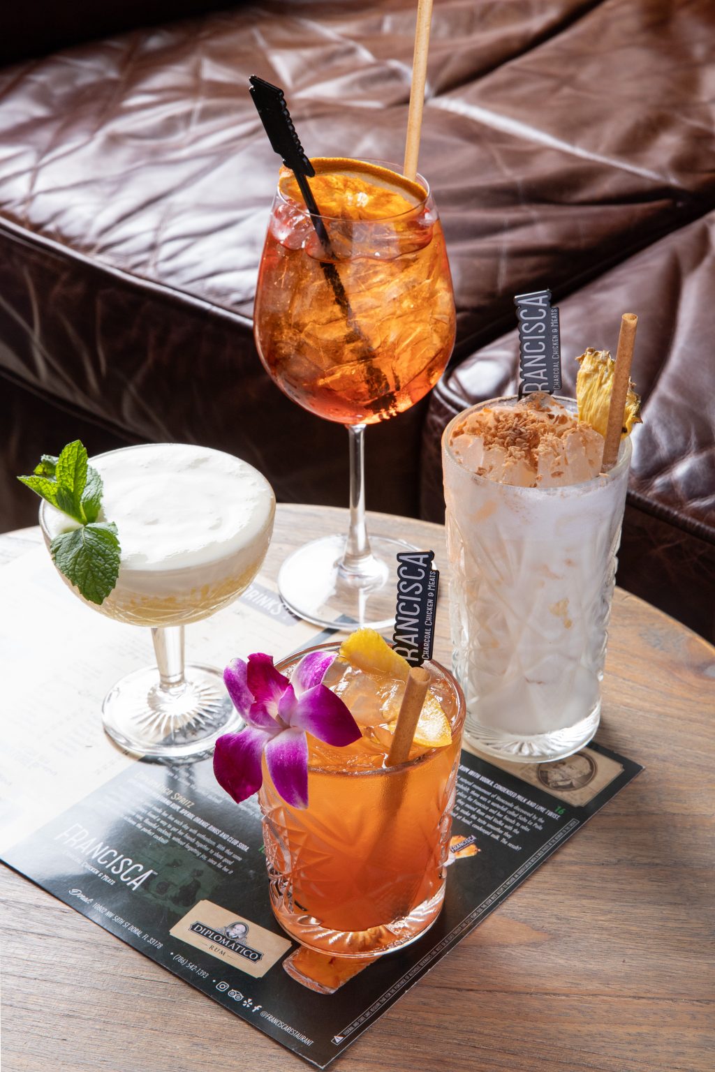 A group of four colorful cocktails arranged on a small round table. The drinks include a mint-garnished frothy cocktail in a coupe glass, an orange spritz in a tall wine glass, a creamy cocktail topped with toasted coconut in a highball glass, and a bright orange drink garnished with an orchid and citrus peel in a rocks glass. A Francisca-branded drink menu lies beneath the glasses.