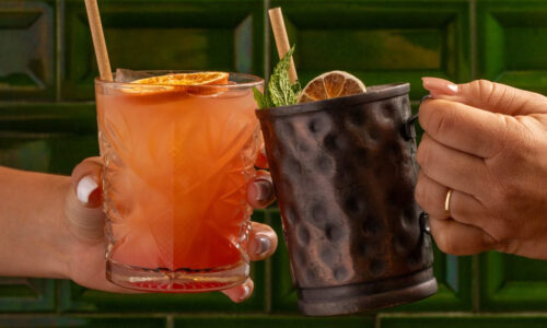 Two hands clink colorful cocktails: one in a crystal glass with citrus slices, the other in a dark hammered metal mug garnished with mint, against a green tiled background.