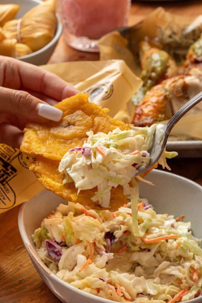 A hand holds a crispy tostone while a spoon scoops creamy coleslaw onto it, with a bowl of coleslaw and assorted dishes in the background.