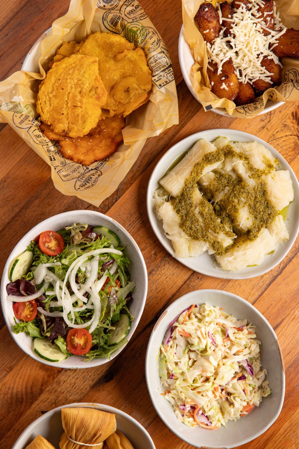 An overhead view of assorted side dishes, including tostones, fried sweet plantains with cheese, yuca with green sauce, a fresh garden salad, creamy coleslaw, and wrapped corn tamales, all arranged on a wooden table.