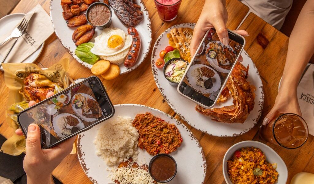 Top-down view of a table filled with assorted Latin dishes like grilled meats, fried fish, rice, beans, plantains, and sides, while two people hold smartphones to photograph the food, surrounded by iced drinks and napkins labeled “Francisca.”