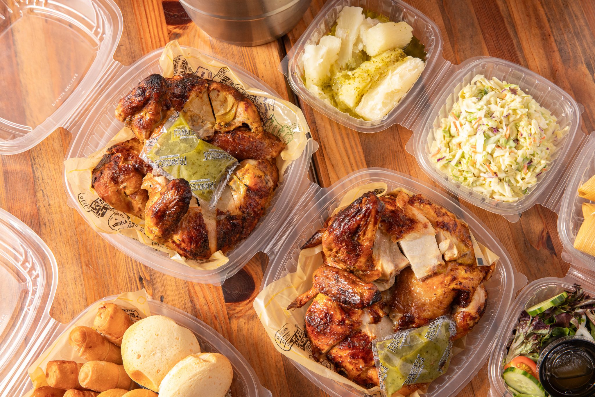 Assorted takeout containers filled with roasted chicken, yuca with sauce, creamy coleslaw, salad, and assorted Venezuelan sides arranged on a wooden table.