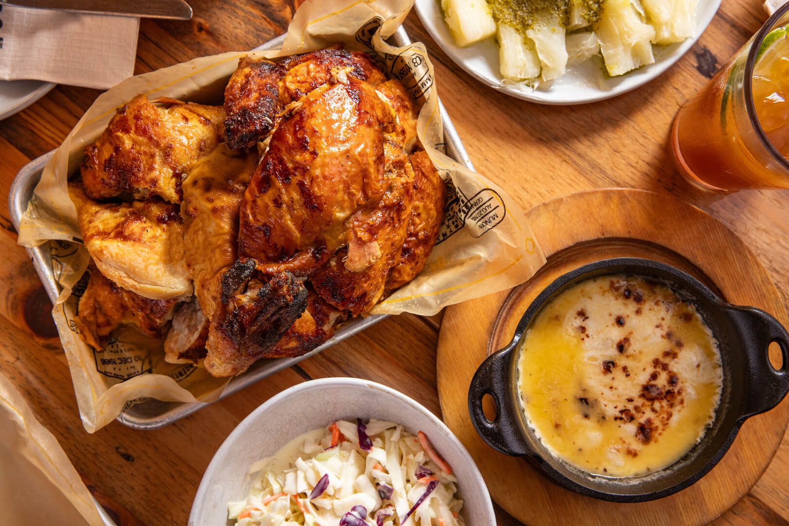A top-down view of a meal featuring a tray of roasted charcoal chicken with crispy golden skin, a small cast-iron dish of melted cheese dip with toasted spots, a bowl of creamy coleslaw, and a side plate of yuca topped with green sauce. A glass of iced tea sits to the right on a wooden table.