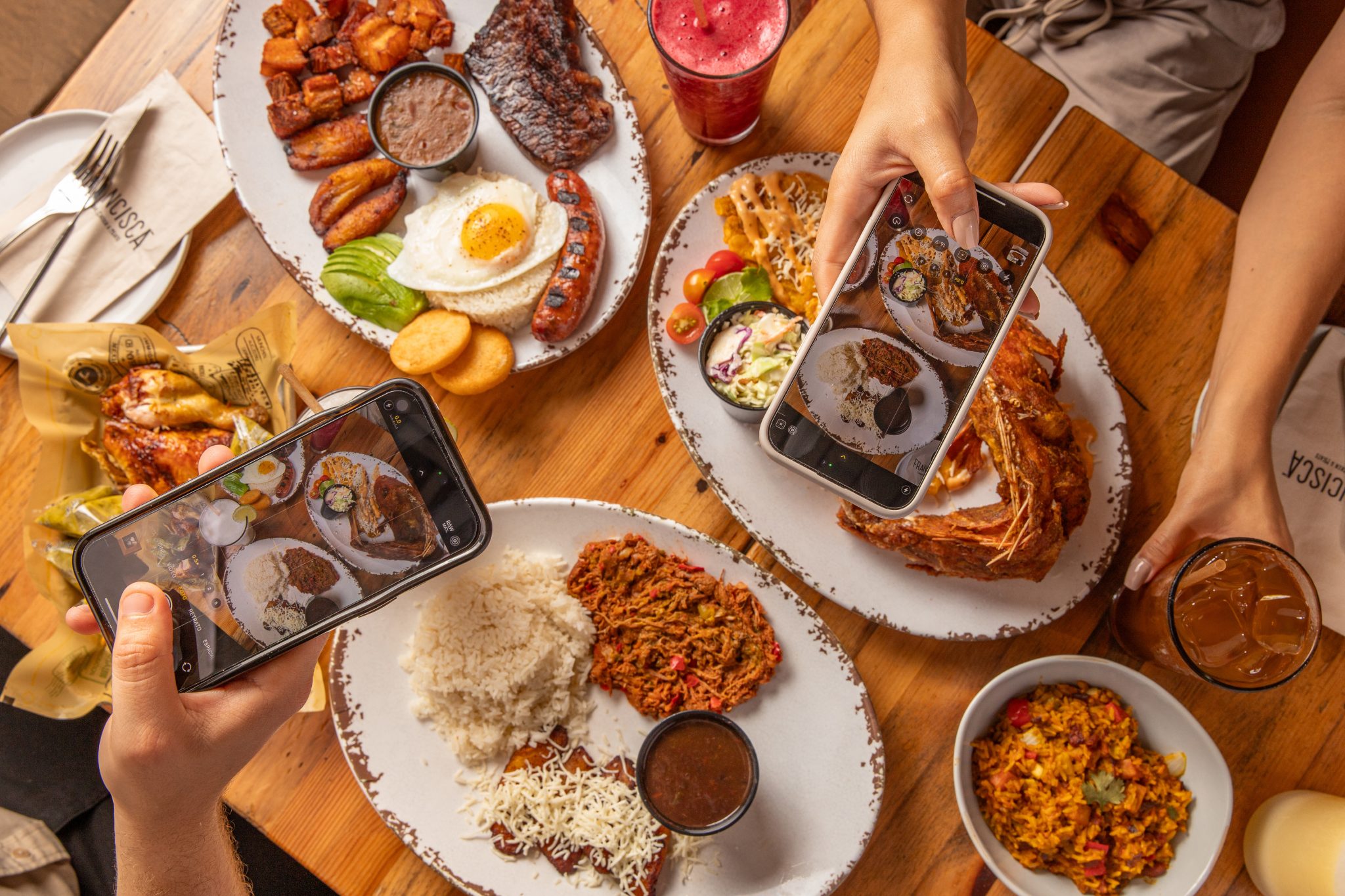 Top-down view of a table filled with assorted Latin dishes like grilled meats, fried fish, rice, beans, plantains, and sides, while two people hold smartphones to photograph the food, surrounded by iced drinks and napkins labeled “Francisca.”