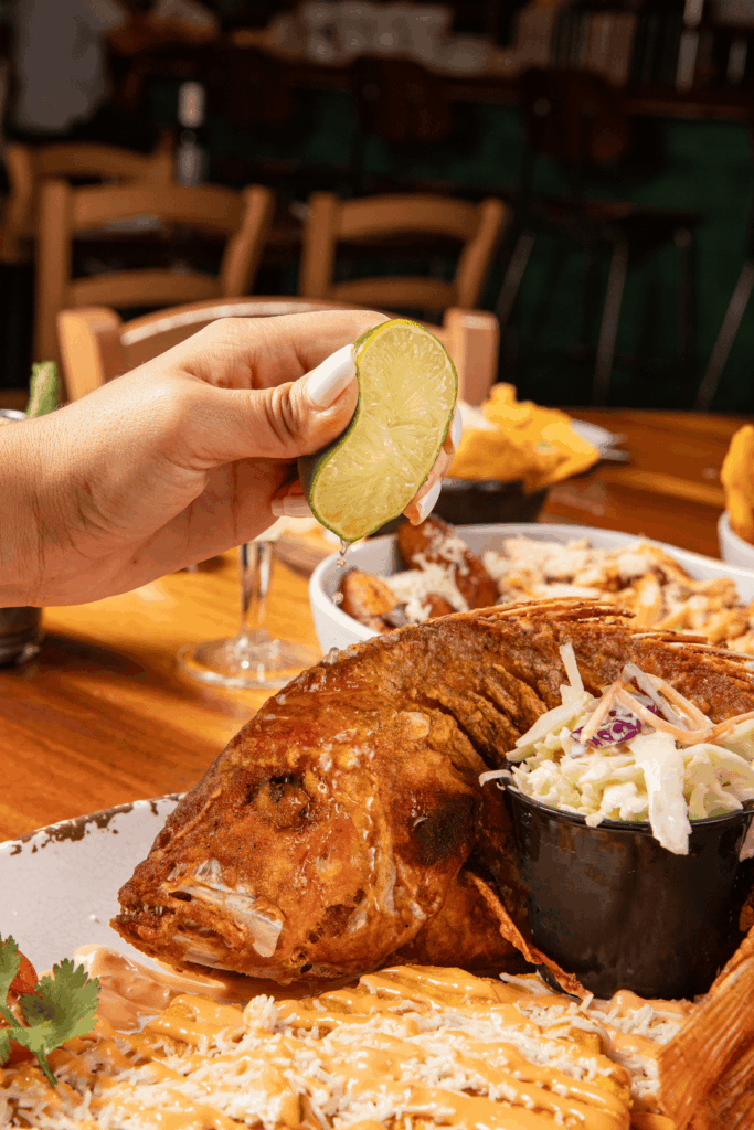 Hand squeezing a fresh lime wedge over a whole fried fish served with coleslaw and a side of cheesy, sauced rice on a restaurant table.