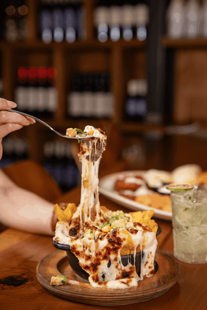 A hand lifts a spoonful of melted, stretchy cheese mixed with toppings from a skillet filled with bubbling, golden-browned queso, garnished with avocado and served with tortilla chips on a wooden board.
