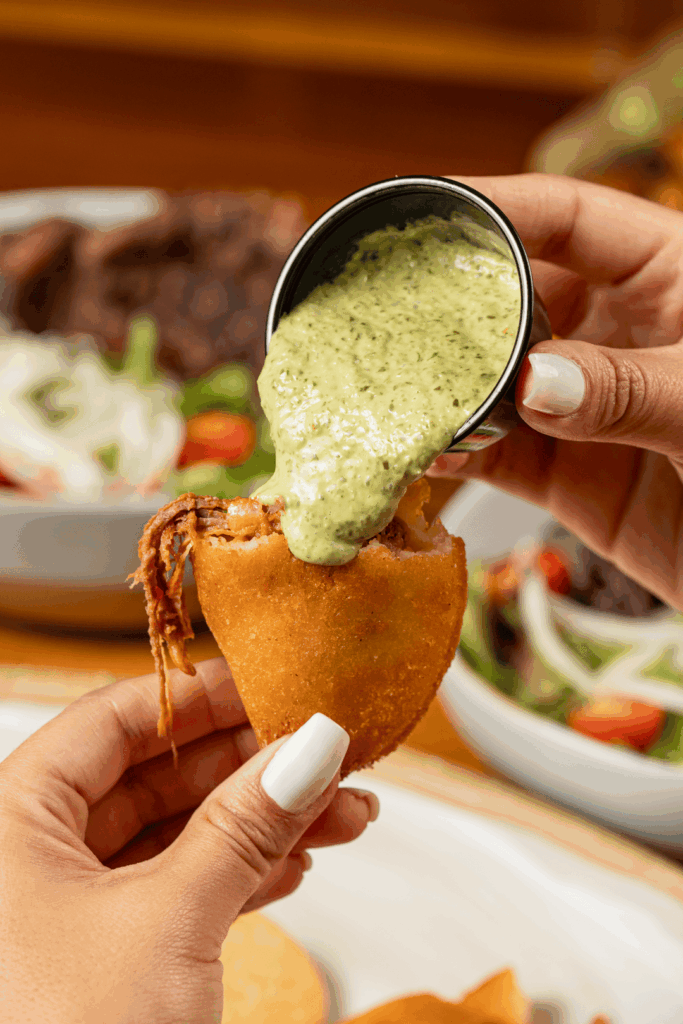 A hand holds a fried empanada filled with shredded meat while another hand pours a creamy green sauce from a small cup over the top.