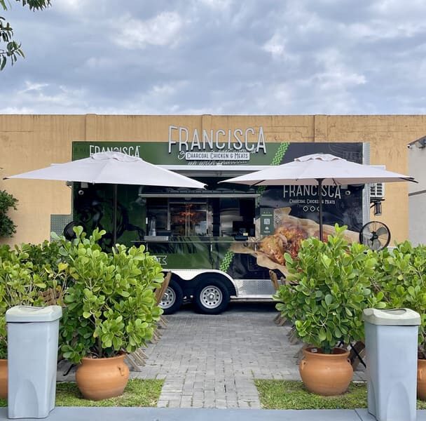 Green Francisca food truck with umbrellas set up in front, surrounded by potted plants and greenery under a cloudy sky.
