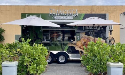 Green Francisca food truck with umbrellas set up in front, surrounded by potted plants and greenery under a cloudy sky.