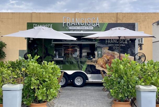 Green Francisca food truck with umbrellas set up in front, surrounded by potted plants and greenery under a cloudy sky.