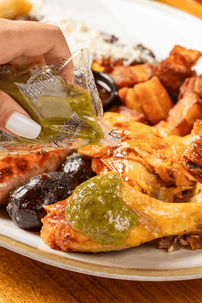 A hand squeezes a packet of green sauce over a serving of roasted chicken, with sausages, crispy pork pieces, and rice visible on the plate.
