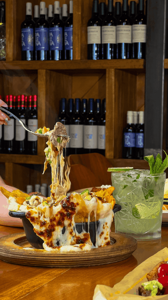 Close-up of a fork lifting a cheesy, loaded nacho bite from a sizzling skillet, with melted cheese stretching dramatically. A refreshing lime and mint cocktail sits beside the dish, with shelves of wine bottles blurred in the background.