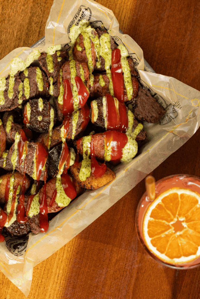 Tray filled with assorted grilled and fried meats drizzled with red sauce and green herb sauce, served on parchment paper with a citrus drink topped with an orange slice beside it.
