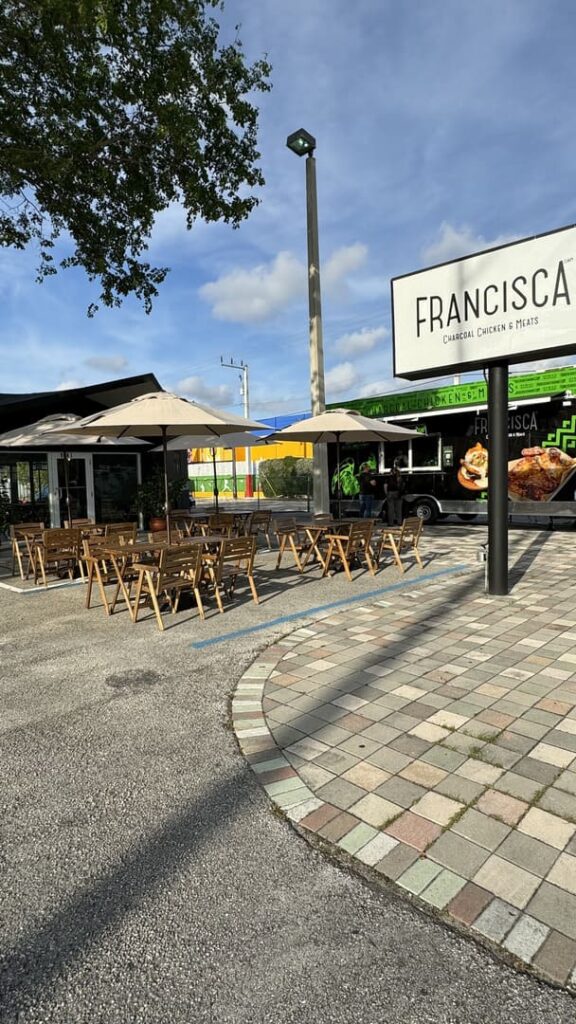 Outdoor dining area with wooden tables and umbrellas in front of a Francisca Charcoal Chicken & Meats food truck, set beside a large branded sign and under a partly cloudy sky.