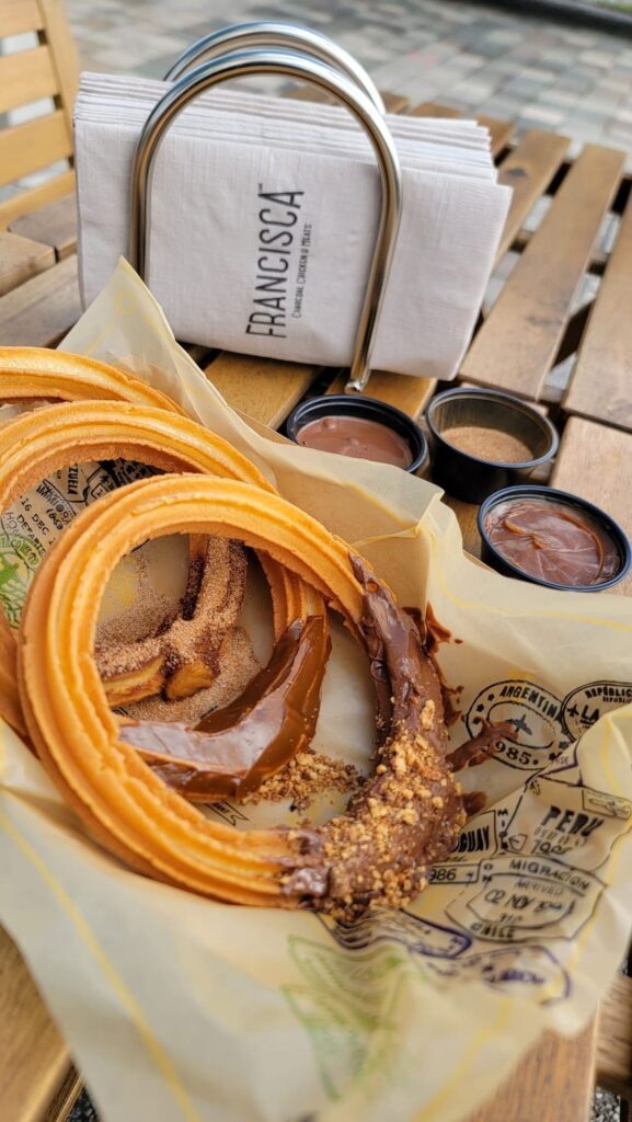 Fresh churros covered in cinnamon sugar and chocolate sauce, served on branded paper beside small dipping cups and a napkin holder labeled “Francisca.”