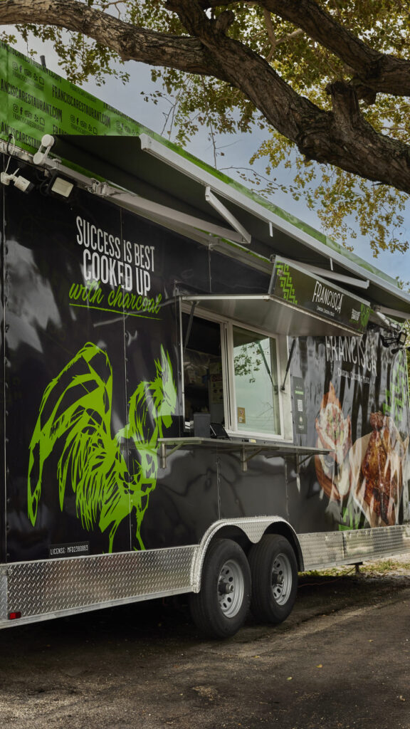Francisca Restaurant’s black and green food truck parked under a large tree, featuring bold graphics of a rooster, grilled chicken imagery, and the phrase “Success is best cooked up with charcoal,” with the serving window open and ready for customers.