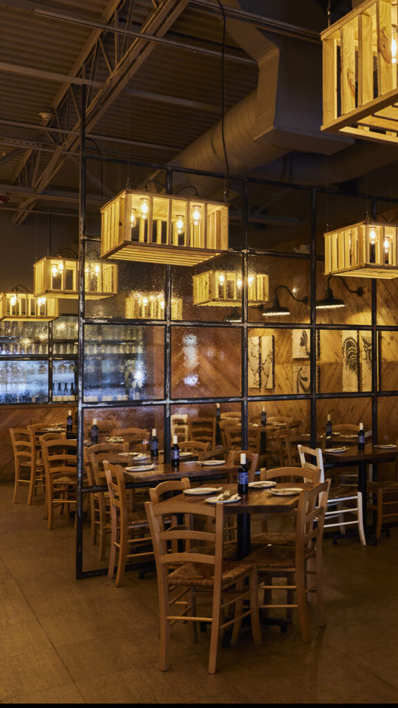 Warm, rustic dining area featuring wooden tables and chairs, dim ambient lighting, crate-style pendant lights, and a large glass partition reflecting the cozy interior décor.