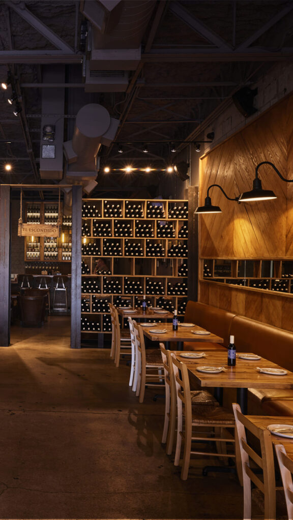 Cozy restaurant interior with wooden tables and chairs, warm wall lighting, and a large shelving unit filled with wine bottles, creating an inviting and rustic dining atmosphere.