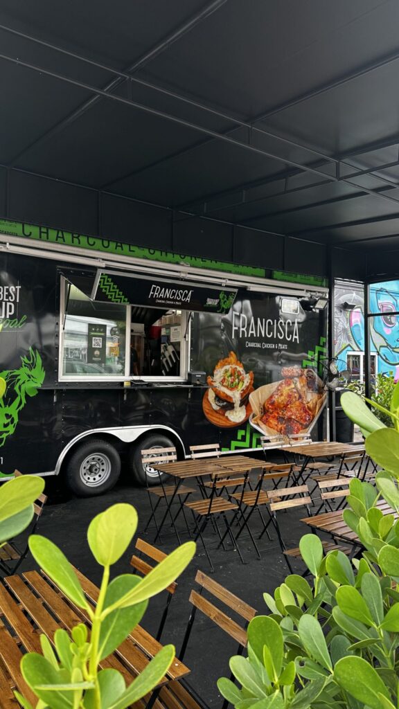 Outdoor dining area with wooden tables and chairs arranged in front of a black Francisca food truck, decorated with images of charcoal-roasted chicken and green branding, partially framed by leafy green plants.