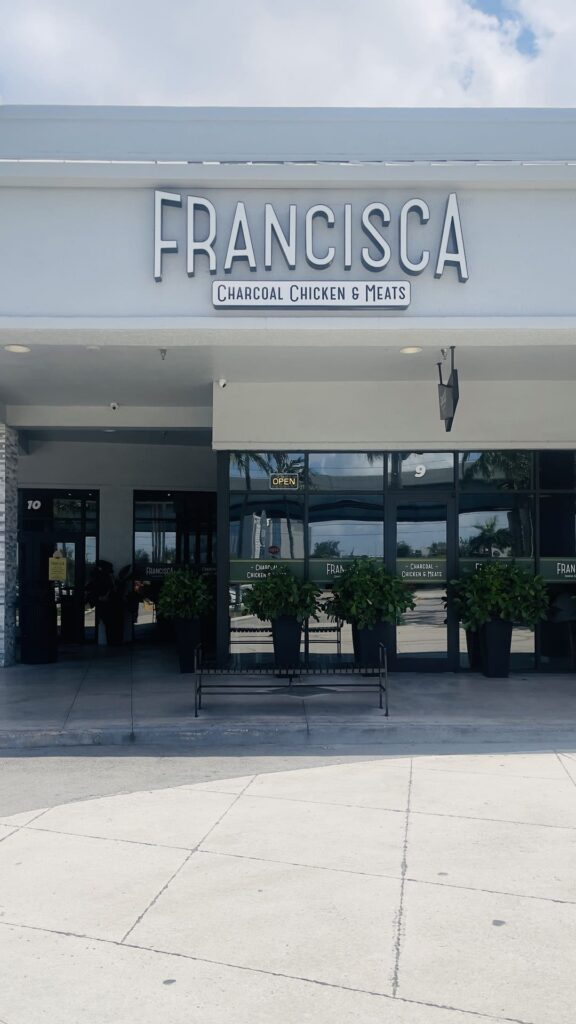 Exterior view of a Francisca Charcoal Chicken & Meats restaurant, featuring a large front sign, potted plants by the entrance, and glass doors reflecting the surroundings on a bright day.