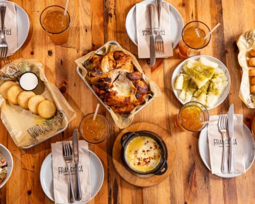 A top-down view of a wooden table set with a family-style spread from Francisca. At the center is a roasted chicken in a tray, surrounded by plates of fried arepas with dipping sauce, yucca topped with green sauce, cheese dip in a small cast-iron pot, tequeños, and a bowl of coleslaw. Several glasses of iced tea and place settings with Francisca-branded napkins, forks, and knives are arranged neatly around the table.