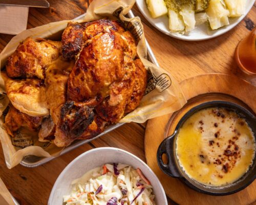 A top-down view of a meal featuring a tray of roasted charcoal chicken with crispy golden skin, a small cast-iron dish of melted cheese dip with toasted spots, a bowl of creamy coleslaw, and a side plate of yuca topped with green sauce. A glass of iced tea sits to the right on a wooden table.