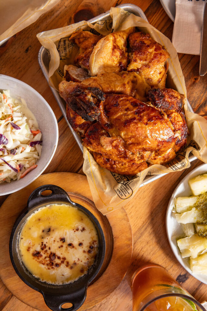 A top-down view of a meal featuring a tray of roasted charcoal chicken with crispy golden skin, a small cast-iron dish of melted cheese dip with toasted spots, a bowl of creamy coleslaw, and a side plate of yuca topped with green sauce. A glass of iced tea sits to the right on a wooden table.