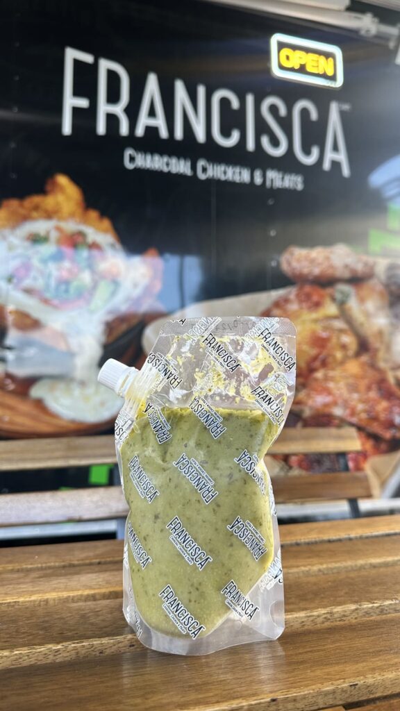 A sealed plastic pouch filled with green sauce sits on a wooden table in front of a Francisca Charcoal Chicken & Meats sign, with a glowing “OPEN” sign visible above it.