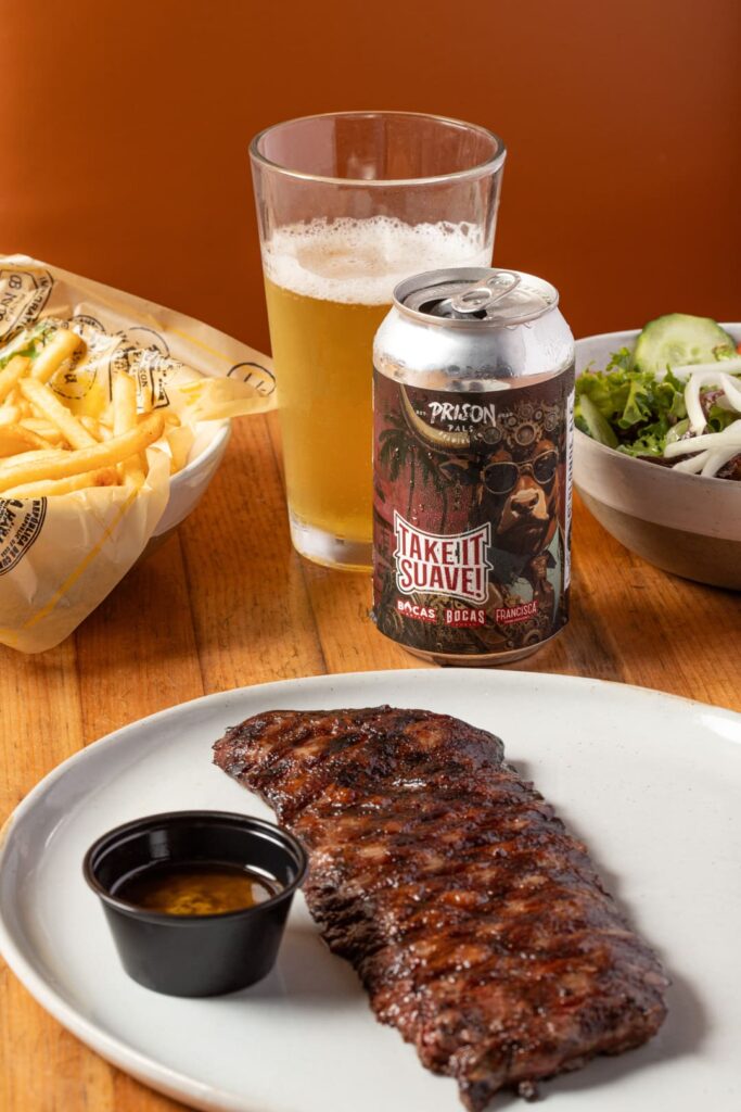 A grilled steak served on a white plate with a small cup of sauce, accompanied by a bowl of fries, a fresh green salad, and a can of beer next to a filled pint glass on a wooden table.
