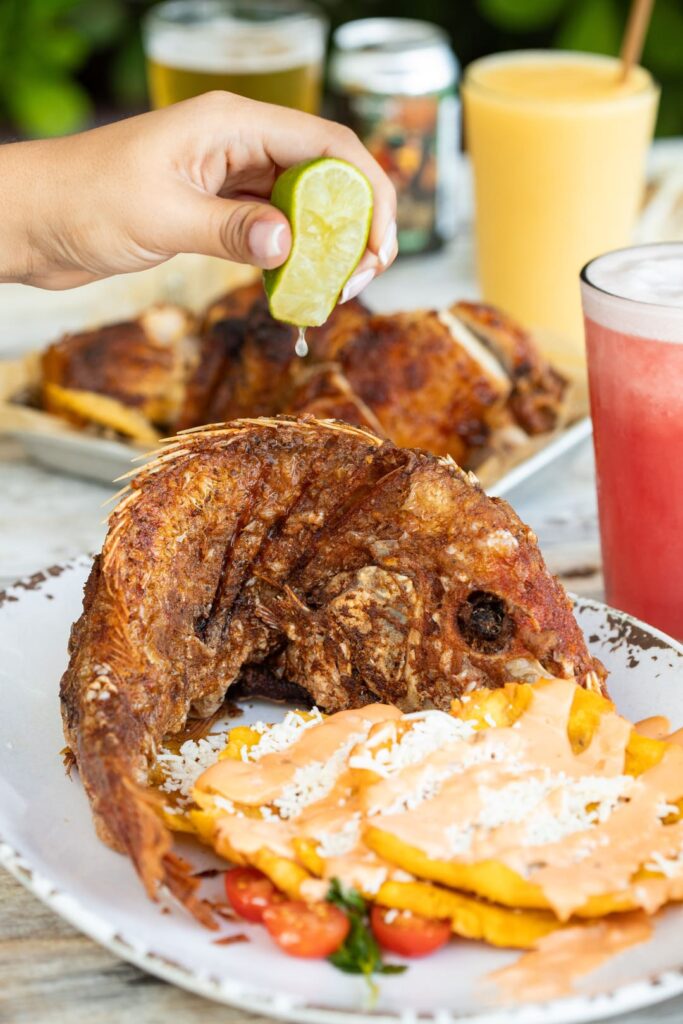 A whole deep-fried fish served on a plate with fried plantains topped with sauce and shredded cheese, alongside cherry tomatoes and herbs. A hand squeezes fresh lime juice over the fish. Colorful drinks and roasted chicken are blurred in the background.