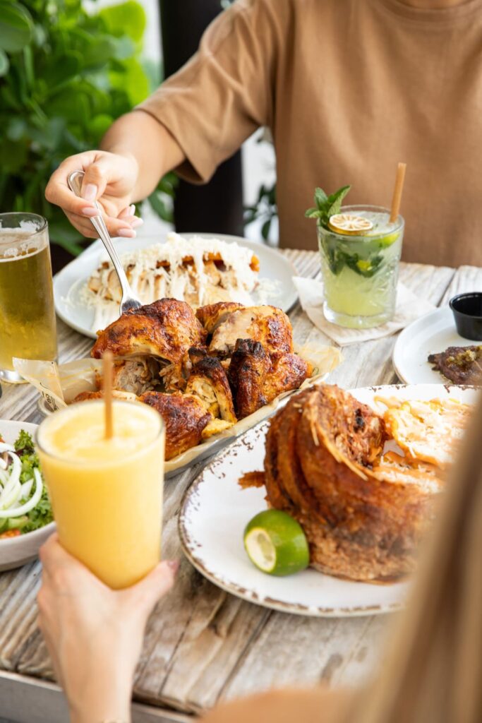 Table scene with several plates of roasted chicken, sides, and drinks. A person in a tan shirt reaches with a fork toward the food while another hand holds a yellow smoothie. A mojito, beer, and additional dishes surround the main platter on a rustic wooden table.