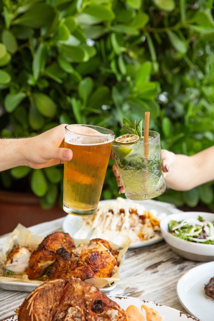 Two people clink glasses: one holding a cold beer, the other a refreshing lime and mint cocktail, over a table filled with roasted chicken and assorted dishes, with lush green foliage in the background.