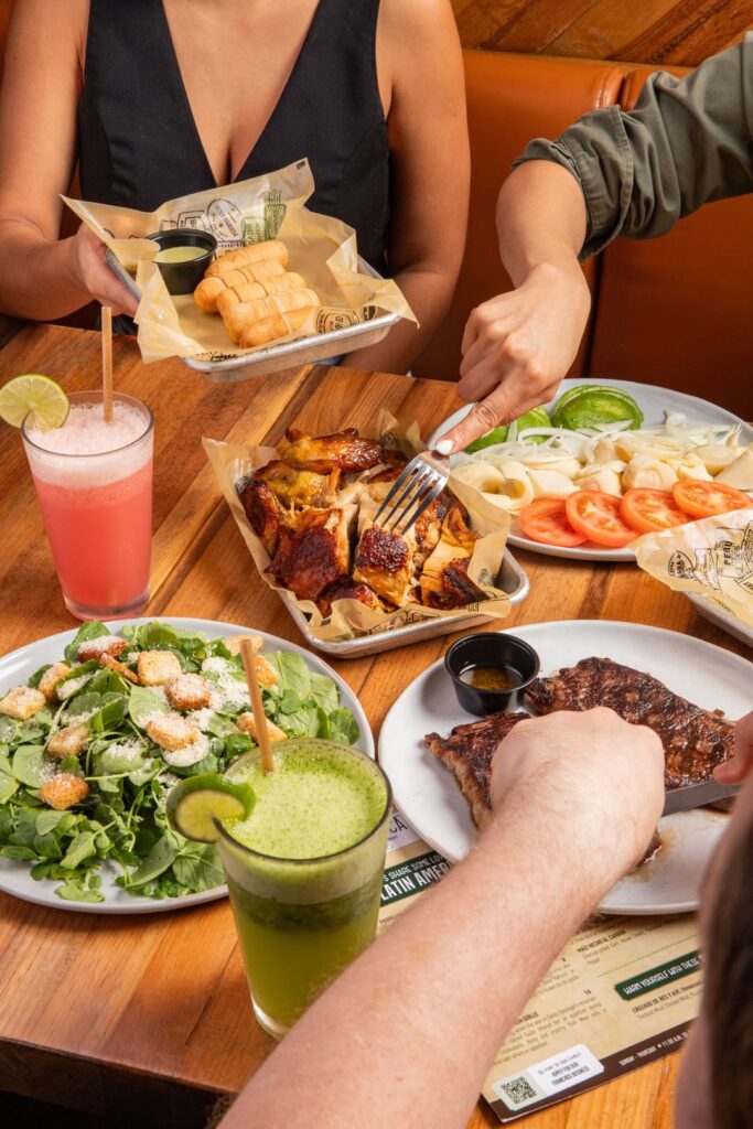 A group of diners share a spread of food at a wooden table, featuring roasted chicken, grilled ribs, fresh salad, heart of palm and tomato slices, tequeños with dipping sauce, and colorful fruit drinks, with hands reaching in to serve and eat.