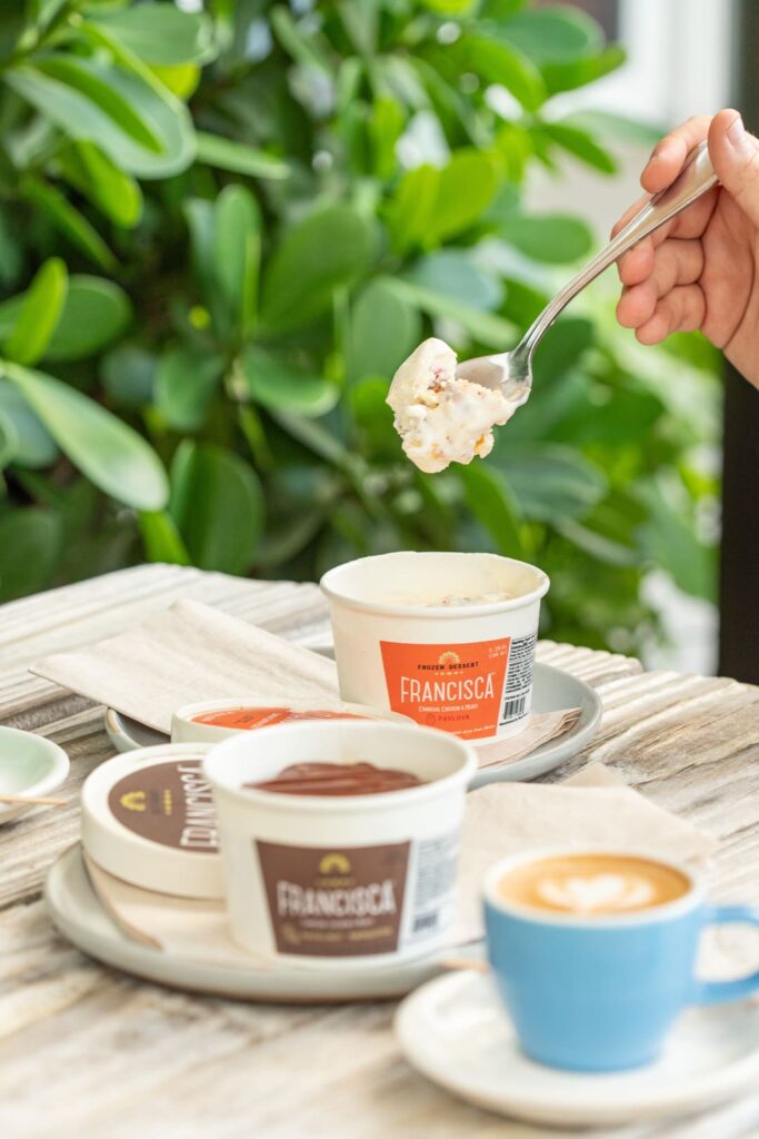 A hand lifts a spoonful of creamy dessert from a Francisca-branded cup on a rustic wooden table, surrounded by other dessert cups and a blue cup of coffee, with green foliage in the background.