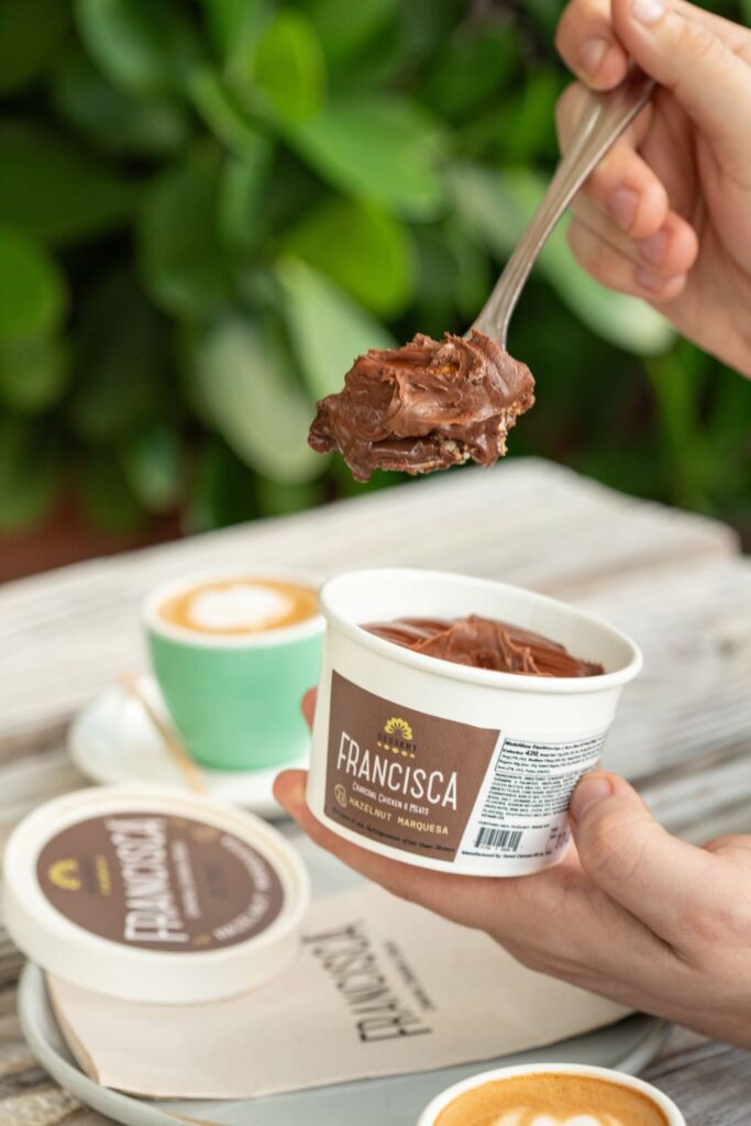 A person scoops a spoonful of rich chocolate hazelnut marquesa from a branded Francisca dessert cup, with the lid placed nearby and two cappuccinos in pastel cups on the table in the background.