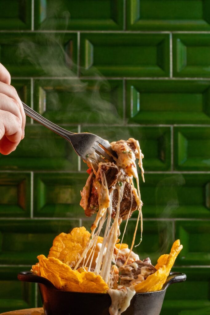 Close-up of a steaming cheesy beef dish being lifted with a fork, with melted cheese stretching above a small cast-iron skillet filled with fried plantains against a green tiled background.