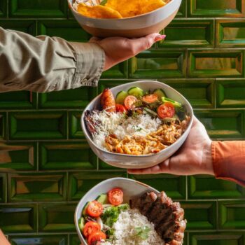 Three people hold three bowls of food stacked vertically against a shiny green tiled wall. The top bowl contains grilled fish, fried plantains, rice, and coleslaw. The middle bowl features shredded chicken, white rice, mixed greens, cucumbers, cherry tomatoes, and fried plantains with grated cheese. The bottom bowl includes grilled beef slices, white rice, mixed greens, cherry tomatoes, cucumber slices, and fried plantains topped with cheese.