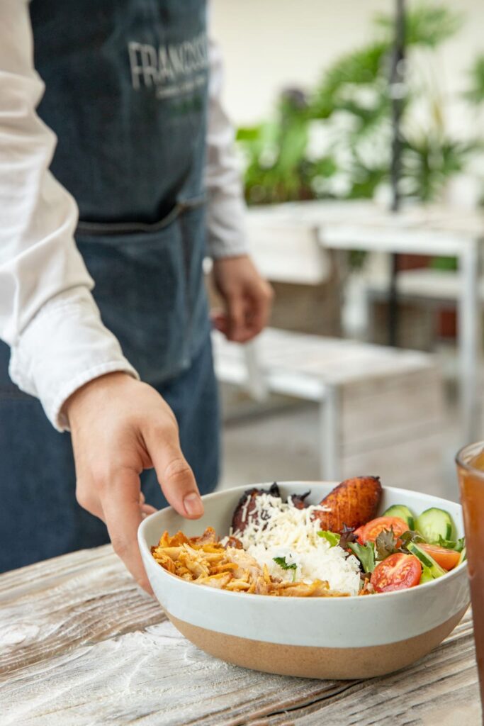 A server wearing a dark apron places a bowl filled with shredded chicken, white rice, fried plantains, fresh greens, cucumbers, and cherry tomatoes on a rustic wooden table.