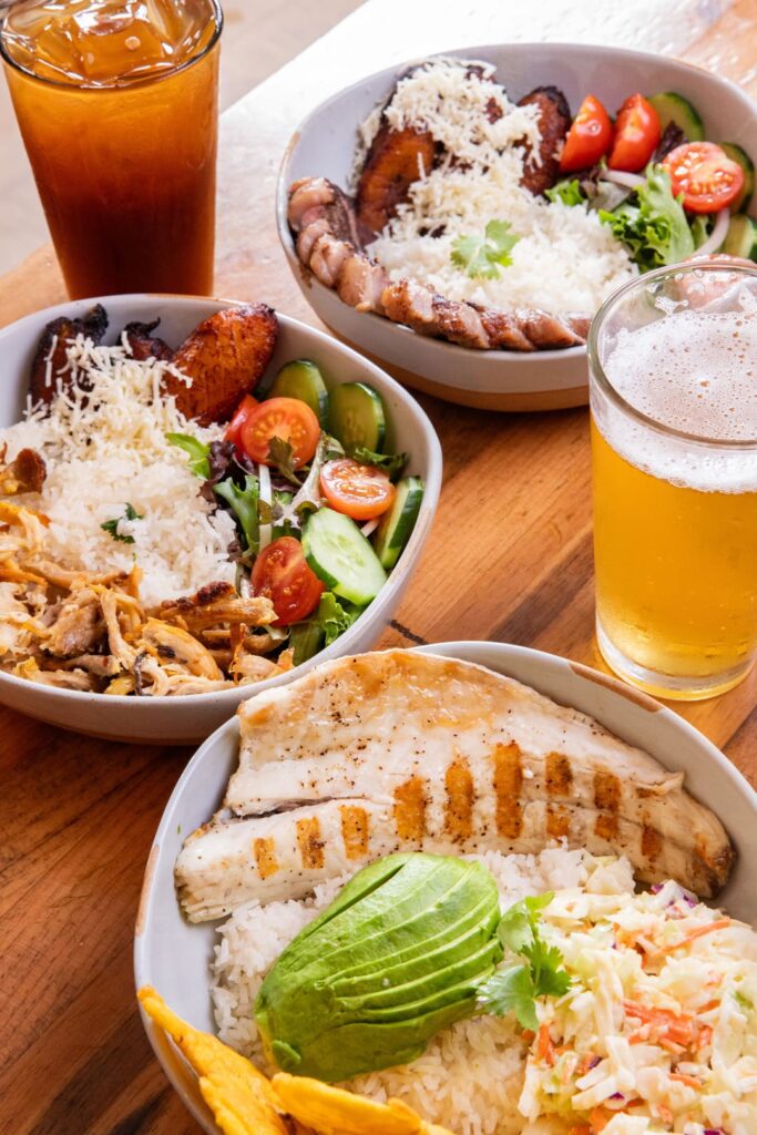 Three bowls filled with rice, fresh salad, plantains, and proteins including shredded chicken, grilled sausage, and grilled fish with avocado, served on a wooden table alongside iced tea and a glass of beer.
