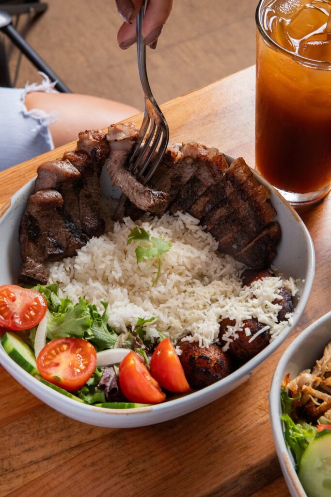 A bowl filled with sliced grilled beef served over white rice, accompanied by fried plantains topped with cheese, mixed greens, cucumber slices, and cherry tomatoes. A person is cutting into the beef with a fork, and a glass of iced tea sits beside the bowl on a wooden table.