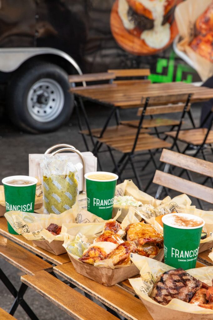 Outdoor table filled with trays of charcoal-roasted chicken, grilled meats, and sides, along with green branded cups and a bag of sauce, with wooden chairs and a food truck visible in the background.