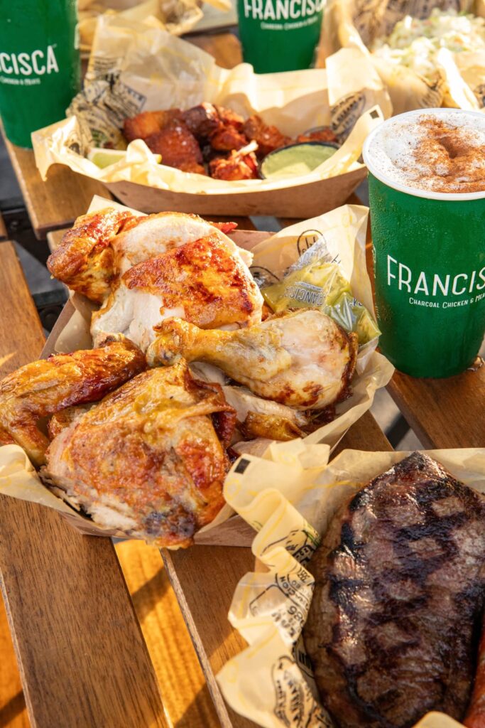 Close-up of takeaway trays filled with roasted chicken pieces, grilled steak, and fried bites, accompanied by green Francisca-branded drink cups on an outdoor wooden table.