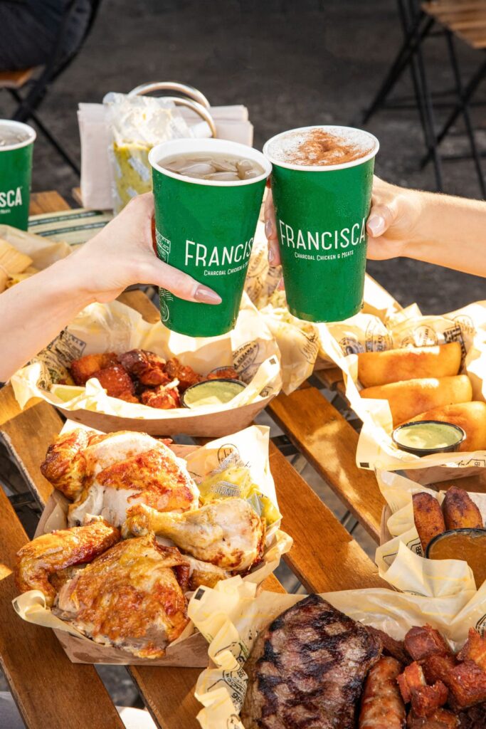 Two people toast with green Francisca-branded cups over a wooden outdoor table filled with trays of roasted chicken, grilled meats, fried bites, and dipping sauces.