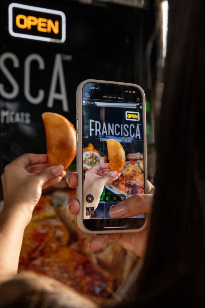 A person holds a fried empanada up to a phone while recording it. The phone screen displays a close-up of the empanada in front of a restaurant sign that reads “Francisca,” with a bright “OPEN” sign glowing above it.