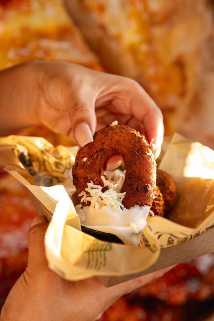 Close-up of a person dipping a crispy, golden-brown fried ring-shaped appetizer into a creamy white sauce, topped with shredded cheese, held in a small paper-lined tray.