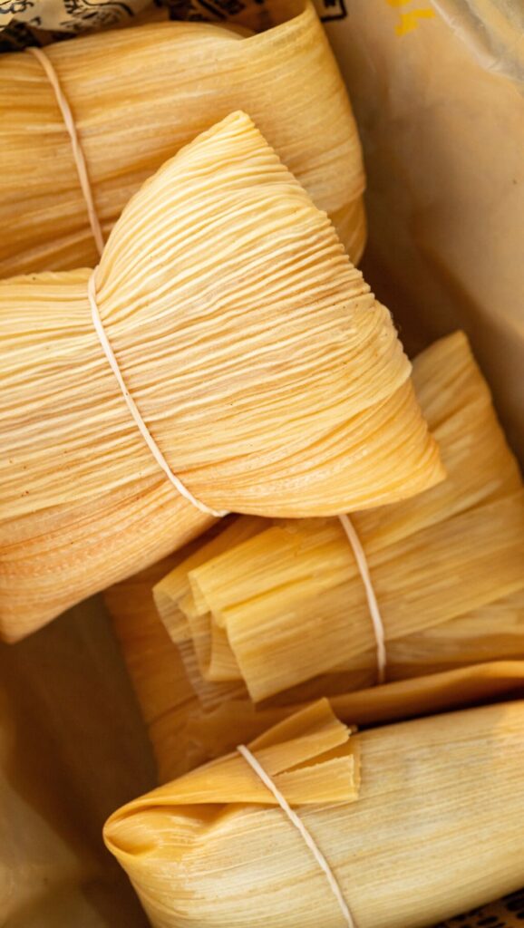Close-up of several wrapped tamales in dried corn husks, tied with thin white strings and arranged inside a container.