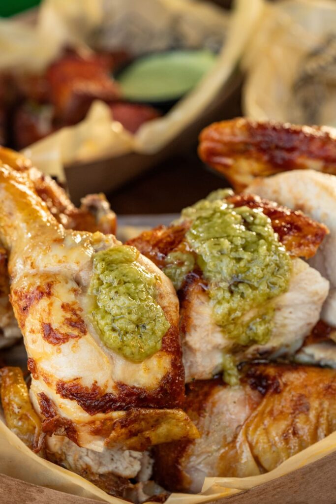 A close-up of juicy, charcoal-roasted chicken pieces topped with a green sauce, served in a paper-lined tray with other dishes blurred in the background.