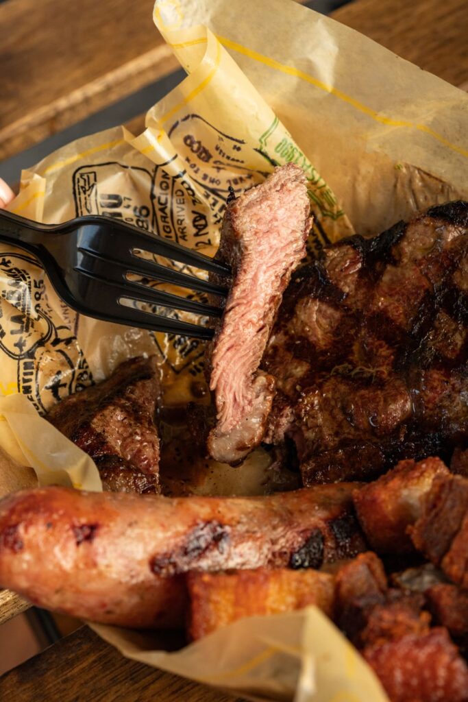 A close-up of a fork lifting a juicy, medium-cooked slice of grilled steak from a tray filled with charbroiled meat, sausage, and crispy pork bites.