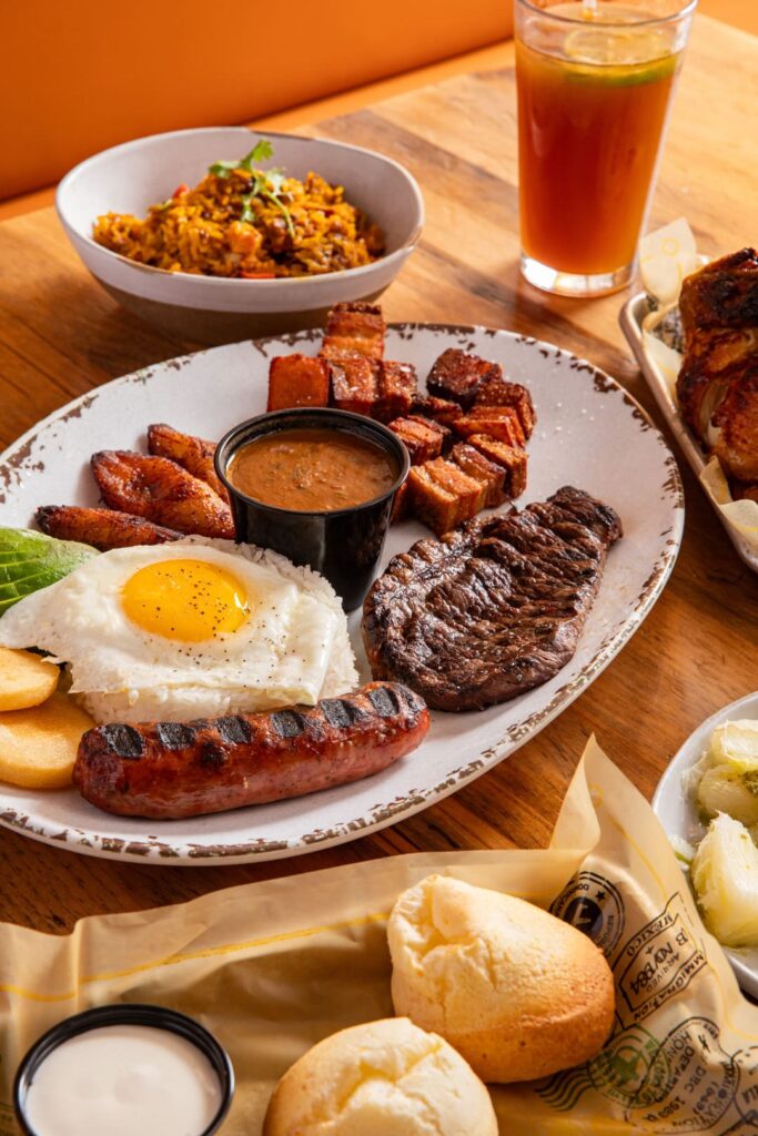 A large plate filled with grilled steak, chorizo, fried egg over rice, fried plantains, avocado slices, and crispy pork belly cubes, served alongside a cup of beans. In the background, there is a bowl of seasoned rice and a glass of iced tea, with small cheese breads on the side.