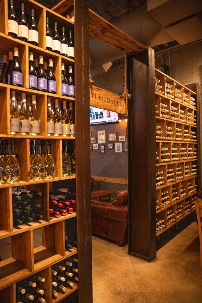 Warm, dimly lit wine cellar room with tall wooden shelves filled with assorted wine bottles. A rustic wooden sign hangs above the entryway, and a cozy seating area with leather chairs is visible in the background.